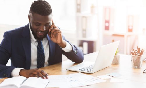 Smiling african american businessman checking marketing research in modern office, talking with assistant by smartphone, empty space